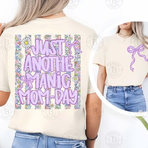May include: Cream-colored t-shirt with the text "JUST ANOTHER MANIC MOM-DAY" in purple bubble letters. The back of the shirt features a floral and striped design. A purple bow is on the front of the shirt.