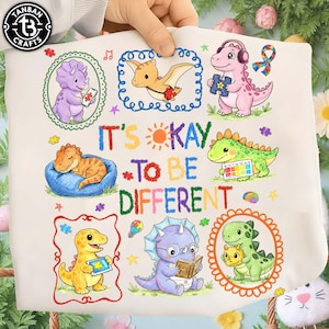 Peut inclure: Tissu blanc avec des dinosaures de dessins animés colorés, des pièces de puzzle et le texte "It's Okay to Be Different". Les dinosaures sont de différentes couleurs et posent, certains portant des écouteurs ou lisant des livres. Le design comprend un soleil et des étoiles.