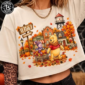 May include: A beige t-shirt featuring a graphic of Winnie the Pooh and Piglet in an autumn town scene. The design includes a wooden sign labeled 47567 Petersburg, a water tower, pumpkins, and fall foliage. The shirt has a crew neck and short sleeves.