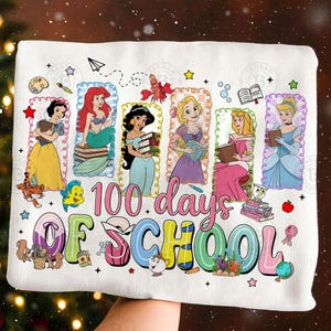 May include: White t-shirt featuring Disney princesses with books, celebrating "100 days of school." The design includes Ariel, Jasmine, Belle, Aurora, and Cinderella, with colorful text and decorative elements.