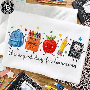 May include: A white t-shirt featuring a colorful graphic of a blue backpack, an orange box of crayons, a red apple, a yellow pencil, and a black and white composition notebook with faces, accompanied by the text "it's a good day for learning".