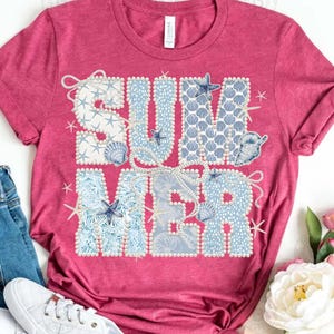 Puede incluir: Una camiseta rosa con la palabra "SUMMER" en un diseño decorativo. Las letras están rellenas de conchas marinas, estrellas de mar y perlas en tonos azules y blancos. La camiseta es de cuello redondo.