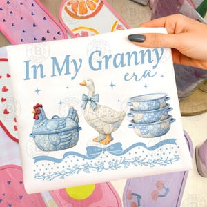 May include: A white decorative sign with the text "In My Granny era." Features a blue chicken, goose, and bowls with floral patterns. The sign is held by a hand, with other decorative items in the background.