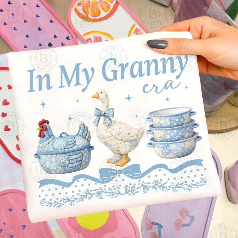 May include: A white decorative sign with the text "In My Granny era." Features a blue chicken, goose, and bowls with floral patterns. The sign is held by a hand, with other decorative items in the background.