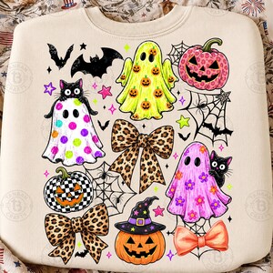 May include: A cream-colored sweatshirt featuring a colorful Halloween-themed graphic print. The design includes ghosts with pumpkin, polka dot, and floral patterns, jack-o'-lanterns, black cats, leopard print bows, bats, and spider webs.