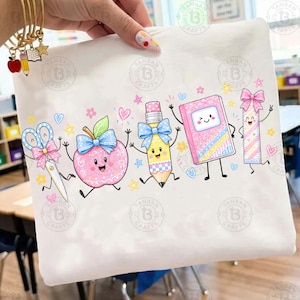 May include: A white t-shirt featuring a colourful graphic of anthropomorphic school supplies including scissors, an apple, a pencil, a notebook, and a ruler, all decorated with blue and pink bows, surrounded by small hearts and stars.
