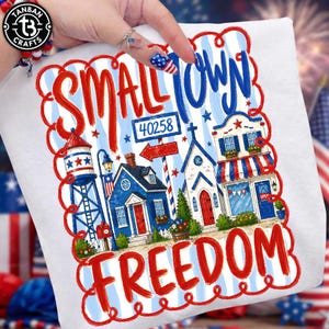 Könnte beinhalten: Ein weißes T-Shirt mit einem Grafikdruck und dem Text Small Town Freedom. Das Design zeigt einen Wasserturm, ein Haus, eine Kirche und ein Geschäft in den Farben Rot, Weiß und Blau sowie ein Schild mit der Zahl 40258.