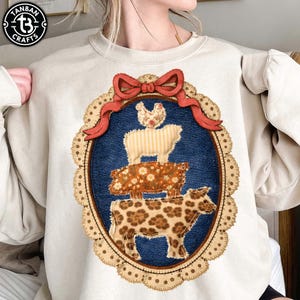 May include: Cream-colored sweatshirt featuring a whimsical farm animal design. The design includes a cow, pig, sheep, and chicken stacked on top of each other within a blue oval frame. A red bow and a scalloped lace border complete the design.