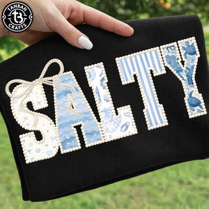 May include: Black towel with the word "SALTY" in a decorative font. The letters are white with blue accents, including stripes, seashells, and turtles. The letter "S" has a bow design. The towel is held by a hand.