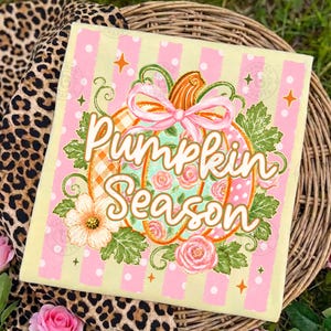 May include: A light yellow t-shirt featuring a graphic of a pumpkin decorated with pink roses and a pink bow. The text Pumpkin Season is printed in white cursive script over the pumpkin. The background has vertical pink and white stripes.