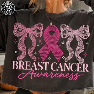 May include: A charcoal gray crewneck sweatshirt featuring a graphic of a pink awareness ribbon flanked by two large pink bows and small star accents. The text "BREAST CANCER Awareness" is printed in pink below the graphic.