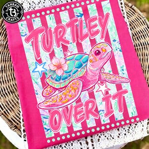 May include: A pink rectangular fabric with a cartoon turtle design. The turtle has a teal shell and a pink body, with a flower on its head. The text "TURTLEY OVER IT" is displayed in pink. The fabric has a striped background.