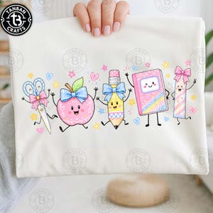 May include: A white t-shirt featuring a colourful graphic of five anthropomorphic school supplies: scissors with a pink bow, a pink apple with a blue bow, a yellow pencil with a blue bow, a pink notebook with a face, and a pink ruler with a pink bow.