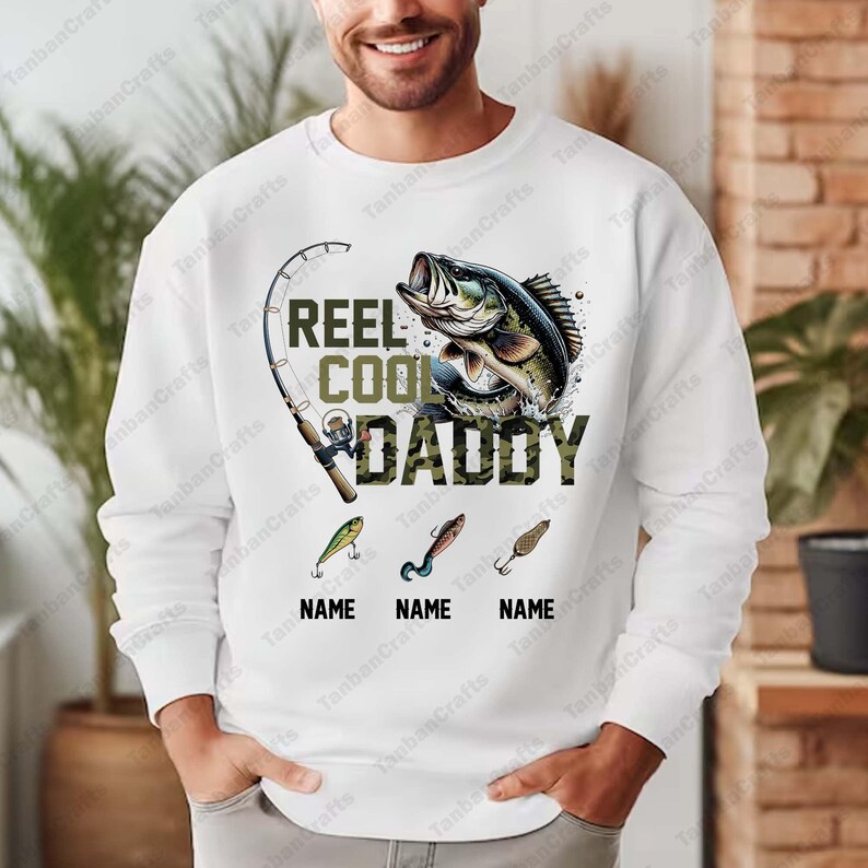 Pu&ograve; includere: Felpa bianca a tema pesca. Presenta una grafica di un pesce che salta, una canna da pesca e la scritta "REEL COOL DADDY". Sotto ci sono tre esche da pesca con spazio per i nomi.