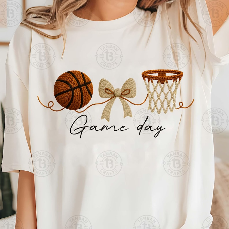 May include: White t-shirt featuring a basketball, a bow, and a basketball net design. The words "Game day" are written in a script font below the graphics. The basketball is brown with black lines.