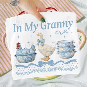 May include: A white decorative kitchen towel with the text "In My Granny era" in light blue. The towel features illustrations of a blue hen, a goose with a blue bow, and three blue bowls.