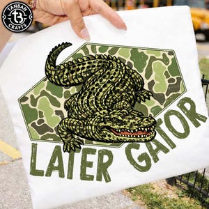 Może przedstawiać: Biała koszulka z szczegółową ilustracją aligatora w odcieniach zieleni i czerni, na tle kamuflażu. Słowa "LATER GATOR" są wydrukowane na zielono pod aligatorem.