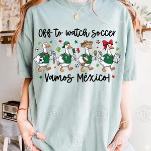 Pu&ograve; includere: Una t-shirt verde chiaro con il testo "Off to watch soccer" e "VAMOS M&Eacute;XICO!" sopra e sotto un grafico di quattro anatre bianche che indossano maglie verdi, che tengono in mano oggetti a tema calcistico come un pallone, una bandiera e un fischietto.