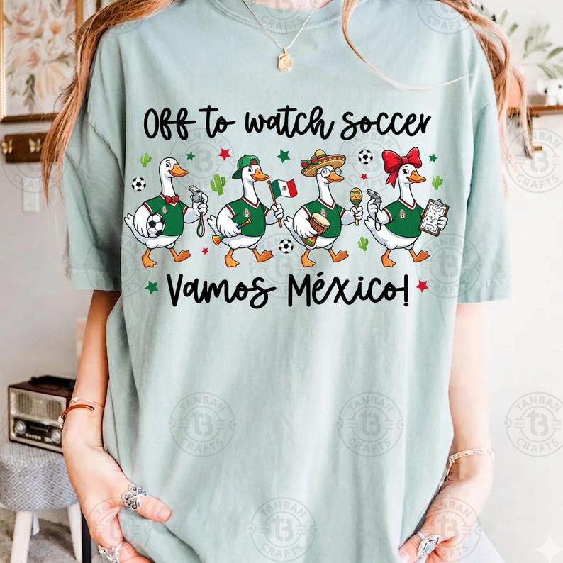 Pu&ograve; includere: Una t-shirt verde chiaro con il testo "Off to watch soccer" e "VAMOS M&Eacute;XICO!" sopra e sotto un grafico di quattro anatre bianche che indossano maglie verdi, che tengono in mano oggetti a tema calcistico come un pallone, una bandiera e un fischietto.