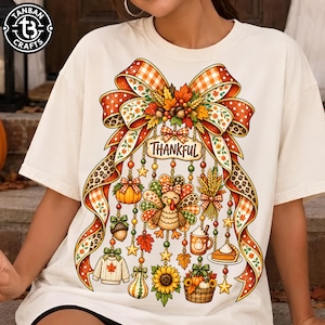 May include: A cream-colored t-shirt featuring a large graphic of a decorative bow with orange, brown, and leopard print patterns. Hanging from the bow are various autumn-themed icons including a turkey, pumpkin, wheat, mug, and the word Thankful.