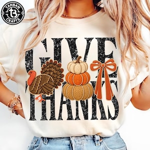 Op de afbeelding: Een crèmekleurig t-shirt met de zwarte, verweerde tekst GIVE THANKS. Het ontwerp bevat een bruine kalkoen, een stapel van drie pompoenen in beige en oranje, en een oranje strik. Een logo in de linkerbovenhoek vermeldt TANBAN CRAFTS.