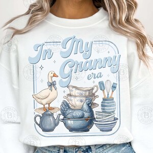 May include: White sweatshirt with a light blue graphic that reads "In My Granny Era." The design includes a goose with a blue bow, a hen, teacups, bowls, and kitchen utensils. The overall theme is vintage and farmhouse.