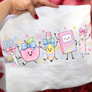 May include: A white t-shirt featuring a colourful graphic print of five anthropomorphic school supplies: scissors with a pink bow, a smiling pink apple with a blue bow, a pencil with a blue bow, a winking pink notebook, and a ruler with a pink bow.