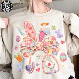 May include: Beige sweatshirt with a colorful Easter-themed design. Features a patchwork bunny bow, stethoscope, Easter eggs, carrots, flowers, and a medical kit. The design includes a syringe, band-aid, and a purple bunny.