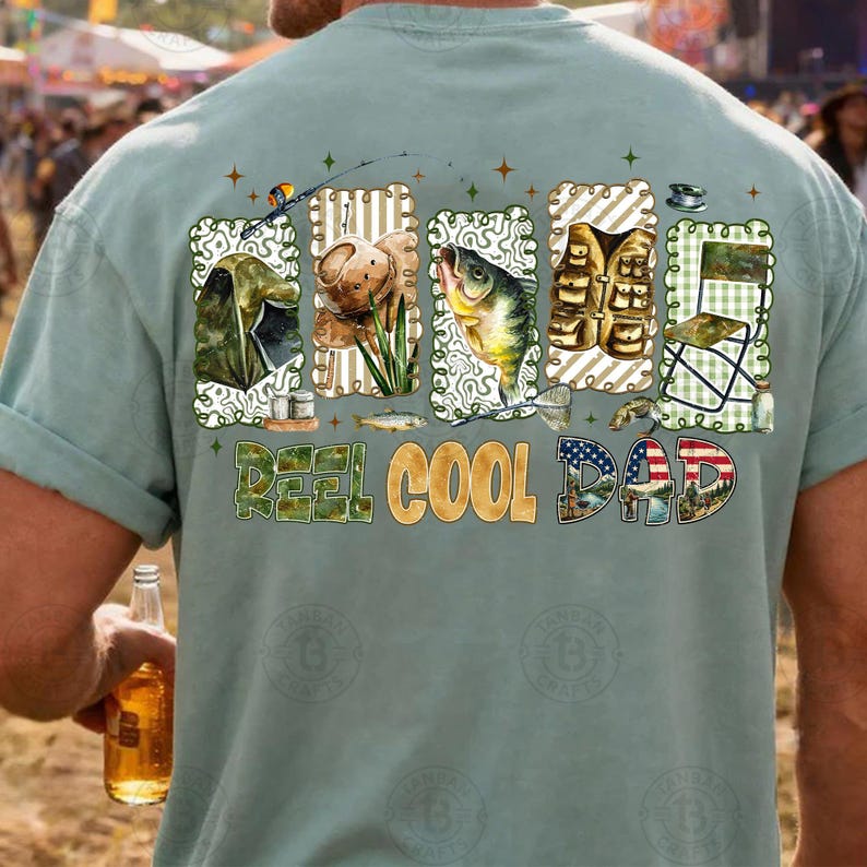 Op de afbeelding: Lichtblauwgroen t-shirt met een visserij-thema grafisch ontwerp. Het ontwerp bevat illustraties van visgerei, een vis en de tekst "REEL COOL DAD" in een mix van groene en gouden letters. De afbeelding bevat ook een Amerikaanse vlag.