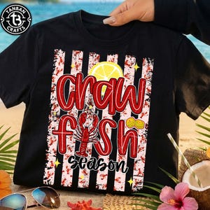 May include: Black t-shirt with a red and white design. The design features the words "Crawfish Season" in red, a red crawfish illustration, and a yellow lemon slice. The background has red and white stripes.
