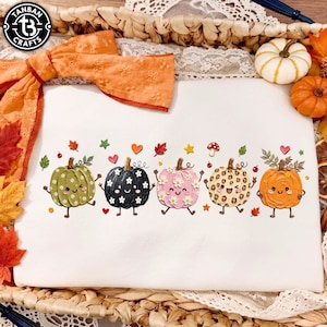 May include: A white sweatshirt featuring a graphic print of five anthropomorphic pumpkins in green, black, pink, leopard print, and orange. Each pumpkin has a smiling face, arms, and legs, surrounded by small colorful leaves, hearts, and stars.