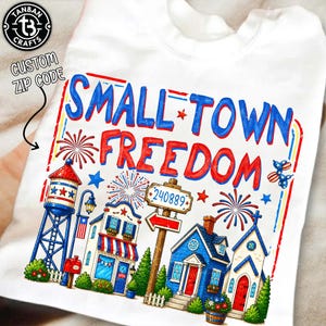 Könnte beinhalten: Weißes T-Shirt mit einem farbenfrohen Grafikdesign. Das Design zeigt die Wörter "SMALL TOWN FREEDOM" in Rot und Blau, zusammen mit Illustrationen eines Wasserturms, Häusern, Feuerwerk und einem Schild mit der Postleitzahl 240889.