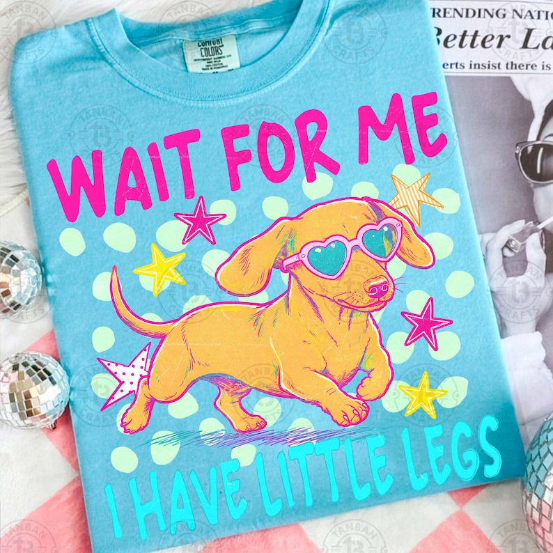 May include: A light blue t-shirt with a cartoon dachshund wearing heart-shaped sunglasses. The shirt has the text "WAIT FOR ME" in pink and "I HAVE LITTLE LEGS" in turquoise, with star and dot graphics.