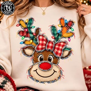 May include: A cream-colored sweatshirt featuring a central graphic of a reindeer face with a red nose, a red and white plaid bow, and antlers decorated with colorful Christmas lights and green plaid fabric patches with frayed edges.