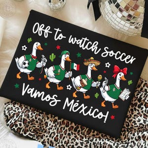 Pu&ograve; includere: Una t-shirt nera con quattro oche in stile cartoon che indossano maglie da calcio verdi. Il testo recita "Off to watch soccer" e "Vamos M&eacute;xico!". Le oche sono decorate con palloni da calcio, una bandiera messicana, un sombrero, una maraca e un fischietto.
