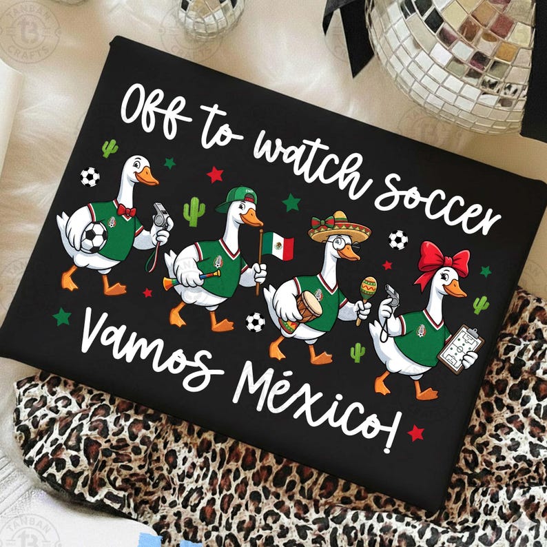 Pu&ograve; includere: Una t-shirt nera con quattro oche in stile cartoon che indossano maglie da calcio verdi. Il testo recita "Off to watch soccer" e "Vamos M&eacute;xico!". Le oche sono decorate con palloni da calcio, una bandiera messicana, un sombrero, una maraca e un fischietto.