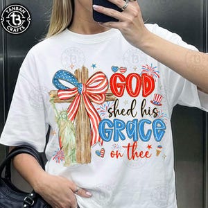 May include: White t-shirt featuring a patriotic design with a cross adorned with a red, white, and blue bow. The shirt includes the text "GOD shed his GRACE on thee" and an illustration of the Statue of Liberty.