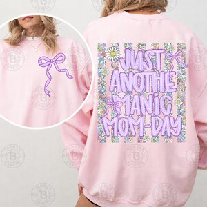 May include: A light pink sweatshirt with a purple bow graphic on the front and back. The back of the sweatshirt features the text "JUST ANOTHER MANIC MOM-DAY" in purple bubble letters, surrounded by a floral design.