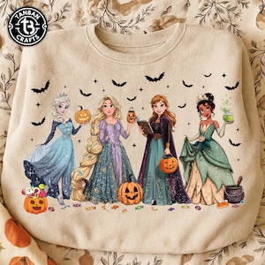 May include: A beige crewneck sweatshirt featuring a graphic print of four animated princesses in Halloween costumes. The design includes pumpkins, bats, stars, and candy scattered around the characters, with a circular logo in the top left corner.