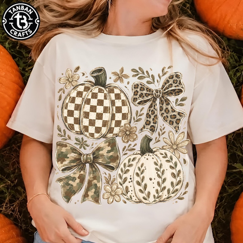 May include: A cream-colored t-shirt featuring a graphic print of two pumpkins and two bows. One pumpkin has a brown and white checkerboard pattern, while the other has a white surface with brown leaf outlines. A leopard print bow and a camouflage bow accompany the pumpkins, along with small floral accents.