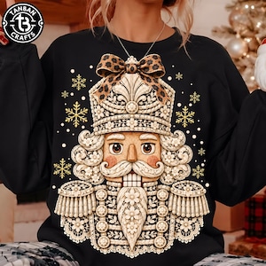May include: A black long-sleeve sweatshirt featuring a large graphic of a nutcracker made of intricate cream-colored lace patterns. The nutcracker wears a leopard print bow on its hat, surrounded by gold-colored snowflakes and white dots.