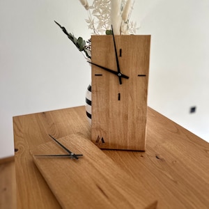 Könnte beinhalten: Zwei Holzuhren mit schwarzen Zeigern und einem schlichten Design. Die Uhren sind aus hellem braunem Holz gefertigt und haben ein kleines Logo auf der Unterseite.
