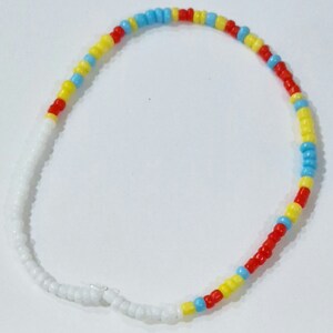 Op de afbeelding: Een witte, rode, gele en blauwe kralen armband. De armband is gemaakt van kleine, ronde kralen die aan elkaar geregen zijn in een herhalend patroon.