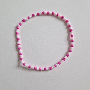 Könnte beinhalten: Ein pinkfarbenes und weißes Armband aus Perlen. Das Armband besteht aus kleinen, runden Perlen, die in einem sich wiederholenden Muster aufgereiht sind.