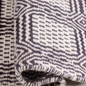 May include: A woven rug featuring a dark charcoal gray and off-white geometric pattern. The textile displays a repeating diamond and linear motif with a thick, textured weave. The edge of the rug is folded over, revealing the dense, knotted construction.