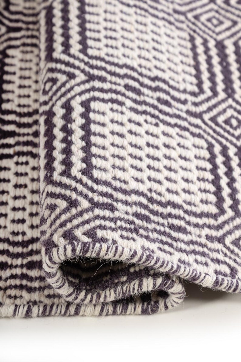 May include: A woven rug featuring a dark charcoal gray and off-white geometric pattern. The textile displays a repeating diamond and linear motif with a thick, textured weave. The edge of the rug is folded over, revealing the dense, knotted construction.