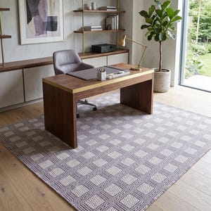 May include: A rectangular area rug featuring a repeating geometric pattern of white squares outlined in dark purple lines. The rug is positioned on a light wood floor beneath a dark wood desk with a gold metal trim and a gray leather desk pad.