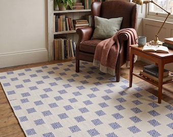 Könnte beinhalten: Ein rechteckiger Teppich mit einem sich wiederholenden Muster aus kleinen, blauen, rautenförmigen geometrischen Motiven auf einem hellcremefarbenen Hintergrund. Der Teppich liegt auf einem Holzboden in einem Raum mit Ledersessel.