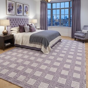 May include: A large rectangular area rug featuring a repeating geometric pattern of light purple and off-white squares. The rug has a textured, woven appearance with a grid-like design that covers the floor in a bedroom setting.
