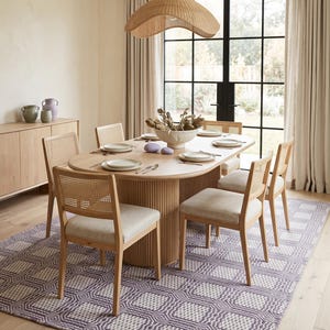May include: An oval light wood dining table with a fluted pedestal base, surrounded by six chairs featuring light wood frames, woven cane backrests, and beige upholstered seats, set upon a purple and white geometric patterned area rug.